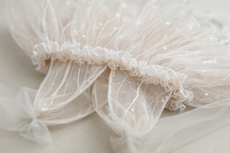 May include: A delicate white lace fabric with a ruffled edge and small sequins. The fabric is sheer and has a soft, flowing texture.
