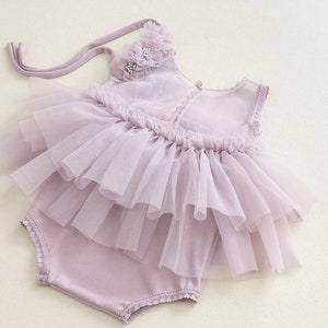 May include: A lavender baby romper with a layered tulle skirt and floral accents. The outfit includes a matching bonnet with a flower detail. The romper is designed with a soft, comfortable fabric and delicate lace trim.