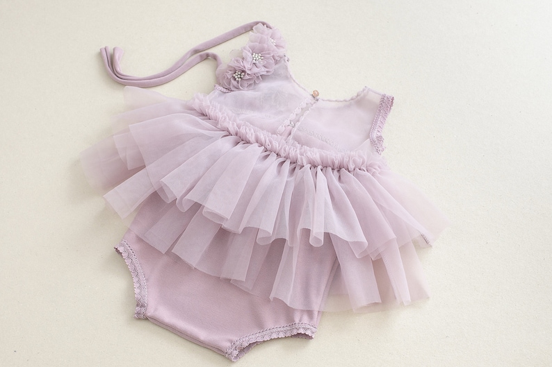 May include: A lavender baby romper with a layered tulle skirt and floral accents. The outfit includes a matching bonnet with a flower detail. The romper is designed with a soft, comfortable fabric and delicate lace trim.