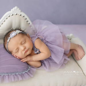 May include: A sleeping baby wearing a lavender tutu dress and a floral headband, resting on a small white couch with a matching lavender pillow. The couch has a decorative back with silver studs.