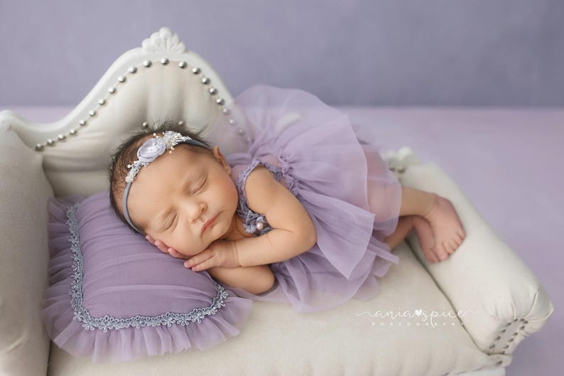 May include: A sleeping baby wearing a lavender tutu dress and a floral headband, resting on a small white couch with a matching lavender pillow. The couch has a decorative back with silver studs.