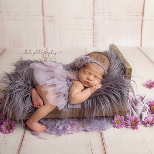 May include: A newborn baby wearing a lavender tutu and headband, sleeping peacefully in a rustic wooden bed filled with gray faux fur. Pink flowers are scattered around the bed, creating a soft, dreamy scene.