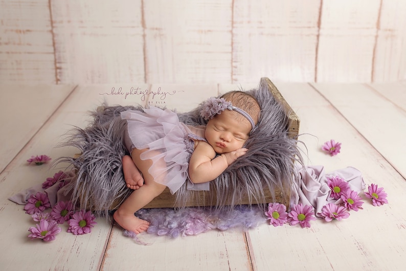 May include: A newborn baby wearing a lavender tutu and headband, sleeping peacefully in a rustic wooden bed filled with gray faux fur. Pink flowers are scattered around the bed, creating a soft, dreamy scene.