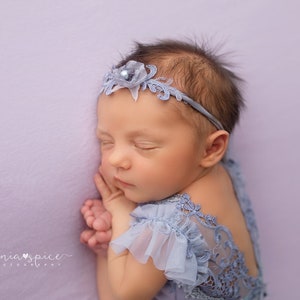 May include: A newborn baby girl sleeping on a lavender background wearing a light blue lace dress and a headband with a flower and pearl embellishment.