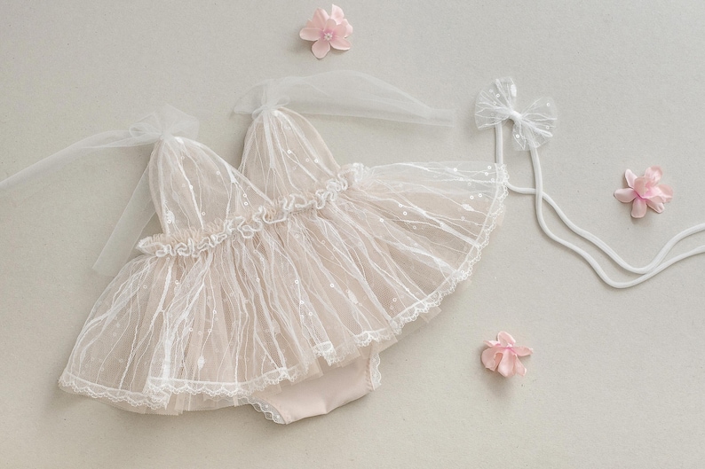May include: A delicate white tulle baby dress with a lace trim and a bow on the back. The dress is laid out on a light gray surface with pink flowers.