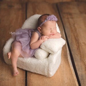 May include: A sleeping infant wearing a lavender tutu and headband rests on a small, white, fluffy chair. The chair is set against a backdrop of wooden planks. The baby is peacefully resting, with arms crossed.