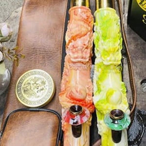 May include: Two decorative smoking pipes with intricate carved designs in orange and green, featuring gold accents. A brown leather carrying case and a gold-colored round container are also visible.