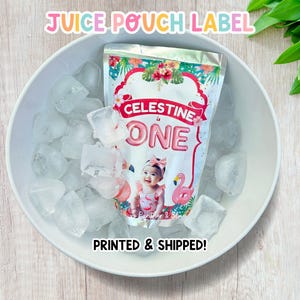 May include: A juice pouch label with the text "CELESTINE is ONE" and a photo of a baby girl. The label has a pink flamingo and tropical flower design. The pouch is in a white bowl filled with ice cubes.