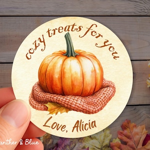 May include: A round sticker with a watercolor illustration of a pumpkin on a knitted blanket. The text "cozy treats for you" and "Love, Alicia" are written in brown script. The sticker has a fall theme.