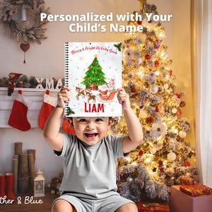 May include: A child holds up a spiral-bound activity book with a Christmas tree, Santa, and reindeer illustration. The book is personalized with the name "LIAM". The background includes a decorated Christmas tree, stockings, and a fireplace.