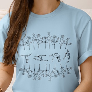May include: Light blue t-shirt with a design of five gymnasts performing different poses, surrounded by a border of stylized flowers.