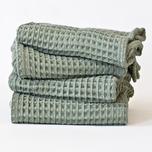 May include: A stack of four sage green waffle weave towels. The towels are folded and stacked on a white surface.