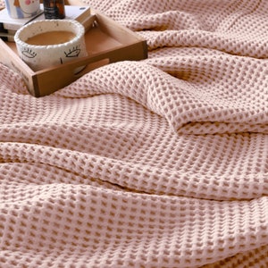 Può includere: Una morbida coperta in tessuto a nido d'ape di colore rosa pallido con un motivo a nido d'ape testurizzato. La coperta è piegata e drappeggiata su un vassoio di legno con una tazza e altri oggetti sopra.