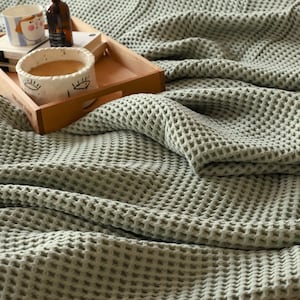 May include: A soft, sage green waffle weave throw blanket with a textured pattern. The blanket is folded and draped over a wooden tray with a mug and a bottle on top.