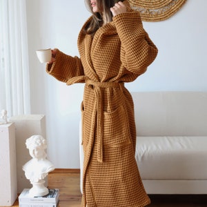 Side view of a camel brown waffle robe with the hood up, model holding a coffee cup by a white sofa – elegant cotton waffle housecoat