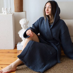 Model sitting on the floor with the hood up in a dark navy waffle robe, classical bust and books beside her – elegant cotton waffle housecoat