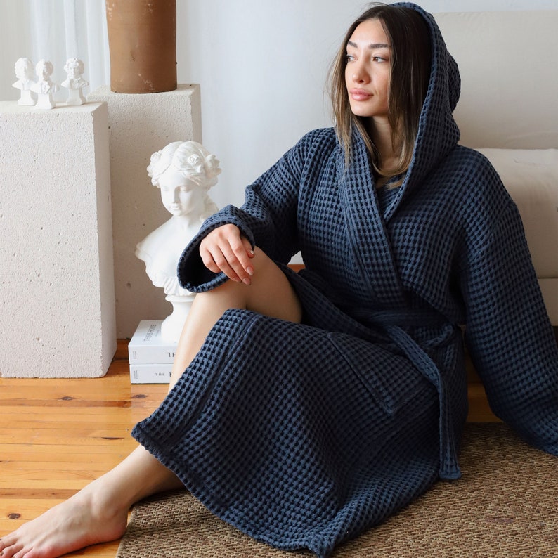 Model sitting on the floor with the hood up in a dark navy waffle robe, classical bust and books beside her – elegant cotton waffle housecoat