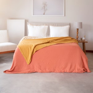 May include: A coral coloured waffle weave throw blanket is draped over a bed with white pillows. A mustard yellow throw blanket is folded on top of the coral blanket.