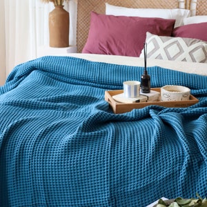 Pode incluir: Uma manta de tecido waffle azul turquesa está drapeada sobre uma cama com almofadas. A manta é texturizada e tem um padrão de waffle. A manta está sobre uma cama com lençóis e almofadas brancas. Há uma bandeja de madeira com uma xícara, uma garrafa e um objeto decorativo sobre a manta.