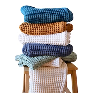 Pode incluir: Uma pilha de toalhas de tecido waffle em várias cores, incluindo azul-petróleo, amarelo mostarda, branco, azul marinho e verde-sálvia. As toalhas estão dobradas e empilhadas em um banquinho de madeira.