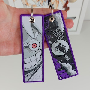 Op de afbeelding: Twee paarse en witte sleutelhangers met een zwart-wit grafisch ontwerp. Een sleutelhanger heeft een schedel met een rood oog en de tekst "HAHA". De andere sleutelhanger heeft een personage met de tekst "HAHA" herhaald.