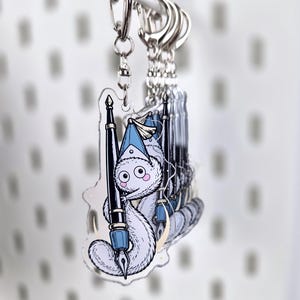 Könnte beinhalten: Ein durchsichtiger Acryl-Schlüsselanhänger mit einer weißen, flauschigen, wurmartigen Kreatur mit großen Augen und rosa Wangen, die einen blauen spitzen Hut trägt. Die Kreatur ist um einen schwarzen und silbernen Füllfederhalter gewickelt.