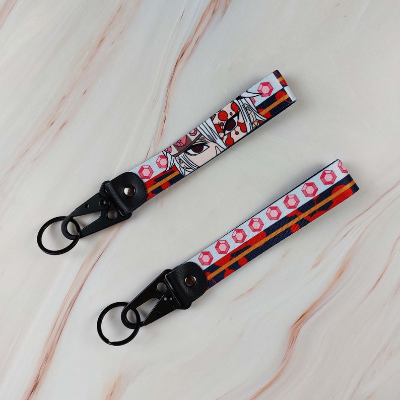 May include: Two fabric wristlet keychains featuring a repeating pattern of red gemstones and a stylized anime character face with white hair and red markings. Each strap is attached to a black metal lobster clasp and a circular split ring.
