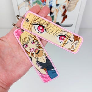 Puede incluir: Dos llaveros rosas y amarillos con ilustraciones de estilo anime. Un llavero presenta un primer plano de la cara de un personaje con ojos rosas y cabello rubio. El otro llavero muestra los ojos de un personaje y parte de su rostro.