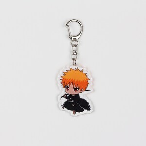 Puede incluir: Un llavero con un personaje de dibujos animados con cabello naranja, vestido con un atuendo negro y sosteniendo una espada. El personaje est&aacute; en estilo chibi, lo que significa que tiene una cabeza grande y un cuerpo peque&ntilde;o. El llavero est&aacute; hecho de acr&iacute;lico transparente.