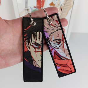 May include: Two black keychains with colorful anime character portraits. The keychain on the left features a man with black hair and red eyes. The keychain on the right features a man with blonde hair and red eyes.