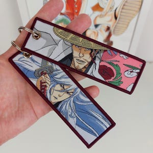 Könnte beinhalten: Zwei Anime-Charakter-Schlüsselanhänger mit burgunderfarbenen Stoffrändern. Der Schlüsselanhänger links zeigt einen Charakter mit weißen Haaren und einem Schwert. Der Schlüsselanhänger rechts zeigt einen Charakter mit einem Strohhut und einem rosa-grünen floralen Hintergrund.