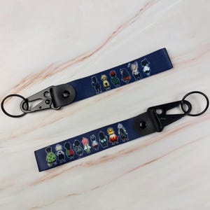 Può includere: Due portachiavi da polso in tessuto blu navy con un motivo ripetuto di piccole illustrazioni colorate di personaggi in stile anime. Ogni portachiavi include un moschettone in metallo nero e un anello fissati da una linguetta in pelle nera.