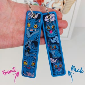Könnte beinhalten: Zwei blaue, bestickte Schlüsselanhänger mit einem Cartoon-Katzen-Design. Der vordere Schlüsselanhänger zeigt eine Katze mit breitem Grinsen, während der hintere Schlüsselanhänger eine Katze mit herausgestreckter Zunge zeigt. Beide Schlüsselanhänger haben einen blauen Rand und einen silbernen Ring.