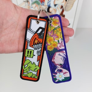 May include: Two rectangular embroidered keychains attached to a metal ring. The left keychain features an orange chainsaw graphic with green Japanese text and a white background. The right keychain displays a purple border with a floral design.