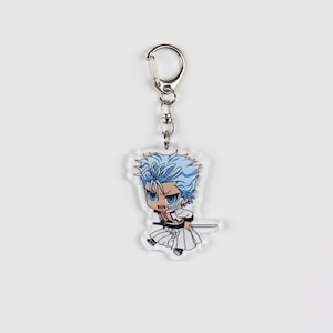 Puede incluir: Un llavero de acr&iacute;lico transparente con un personaje de anime de estilo chibi con cabello azul, un atuendo blanco y negro y una espada. El personaje tiene una expresi&oacute;n seria y sostiene la espada en su mano derecha.