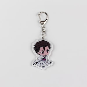 Puede incluir: Un llavero con un personaje de anime blanco y negro con una cadena plateada. El personaje tiene el pelo casta&ntilde;o y lleva una t&uacute;nica blanca con una faja roja.