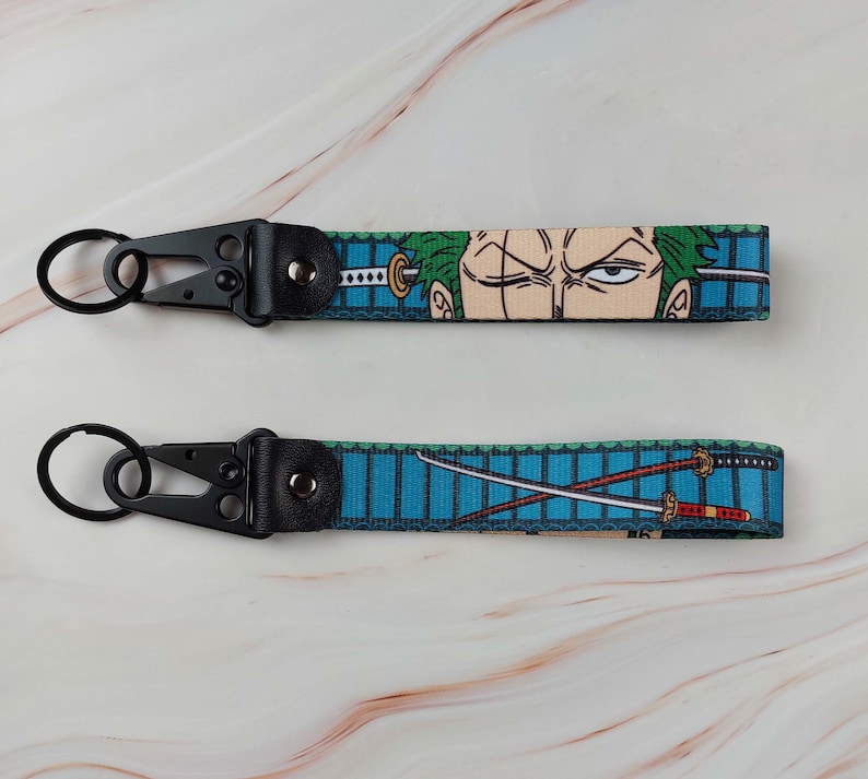 May include: Two fabric wristlet keychains featuring anime-inspired graphics. One strap displays a character with green hair and a sword, while the other shows two crossed swords on a blue background. Both include black metal hardware and a key ring.