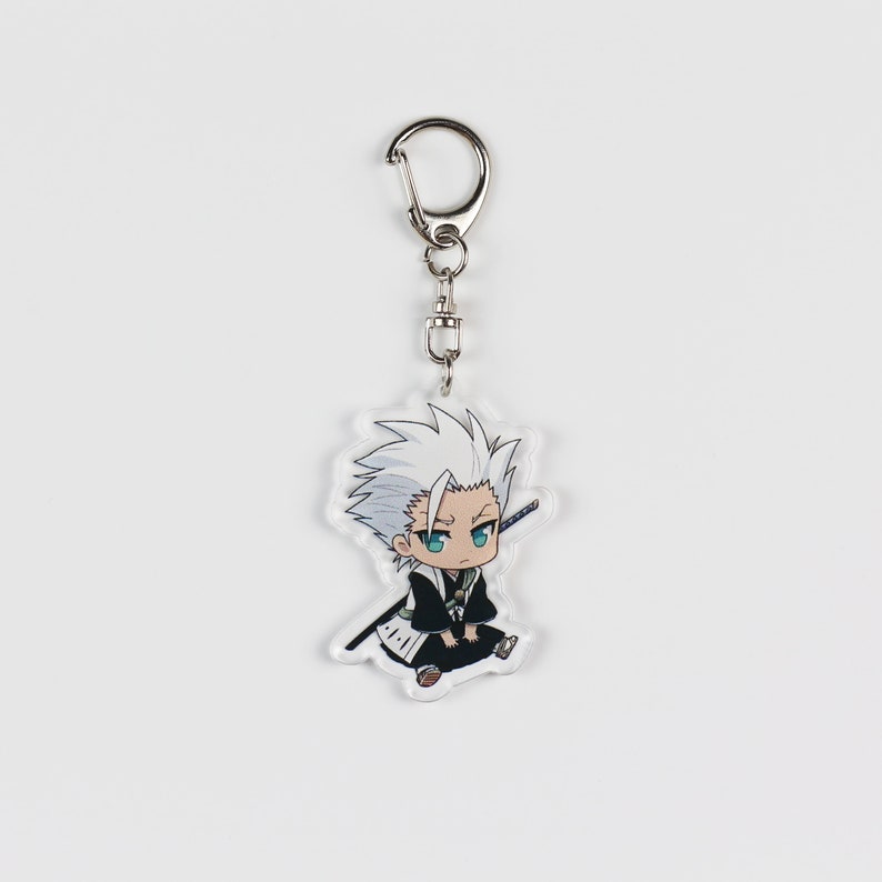 Puede incluir: Un llavero con un personaje de anime blanco y negro con una cadena plateada. El personaje tiene el pelo blanco, los ojos azules y lleva un atuendo blanco y negro.