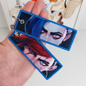 May include: Two blue and black keychains with colourful images of anime characters. The keychain on top features a blue-haired character with red eyes. The keychain on the bottom features a red-haired character with blue eyes. Both keychains have a blue border and the text "VII" on the bottom keychain.