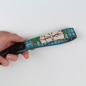 May include: A fabric wrist lanyard featuring a repeating graphic print of a green-haired character with a scar over one eye, holding a sword in their mouth. The strap has a teal grid pattern background and includes a black metal lobster clasp.