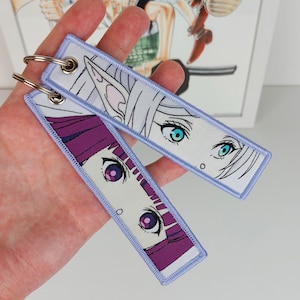 Op de afbeelding: Twee anime-sleutelhangers met paarse en blauwe accenten. De sleutelhangers tonen de gezichten van twee vrouwen met grote ogen. De ene sleutelhanger heeft een witte achtergrond en de andere een paarse achtergrond.