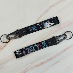 May include: Two black fabric wrist lanyards featuring anime-style graphics. One strap displays the text ARISE with a character illustration, while the other shows a series of small creature figures. Both include black metal lobster clasps and rings.