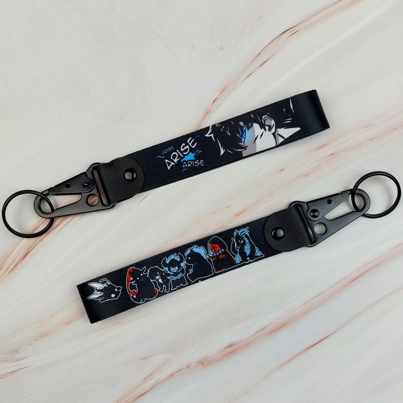 May include: Two black fabric wrist lanyards featuring anime-style graphics. One strap displays the text ARISE with a character illustration, while the other shows a series of small creature figures. Both include black metal lobster clasps and rings.