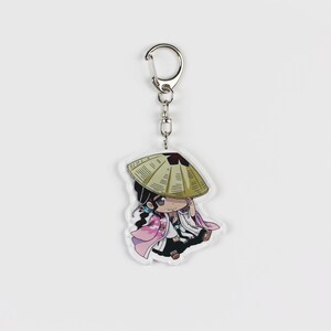 Puede incluir: Un llavero con un peque&ntilde;o personaje de dibujos animados con un sombrero de paja, un kimono rosa y blanco y cabello negro. El llavero est&aacute; unido a un anillo de metal plateado.