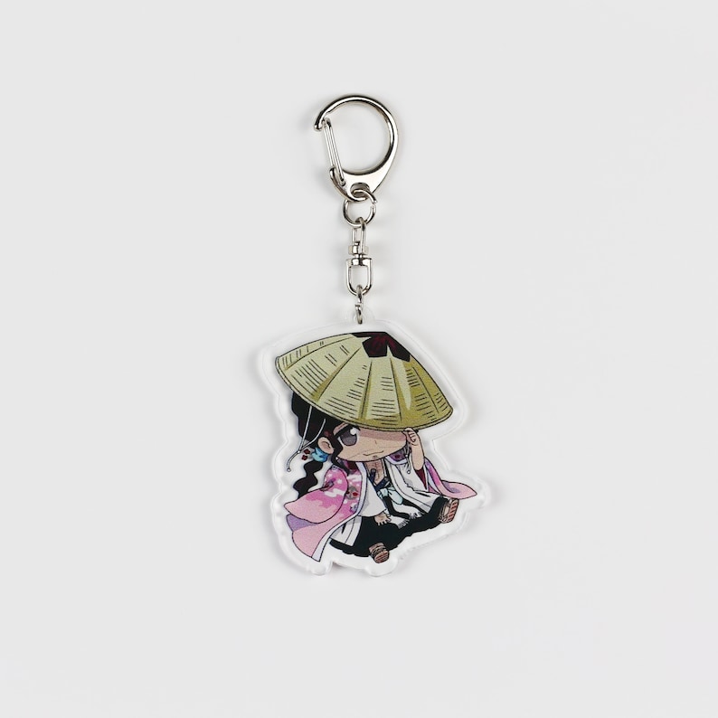 Puede incluir: Un llavero con un peque&ntilde;o personaje de dibujos animados con un sombrero de paja, un kimono rosa y blanco y cabello negro. El llavero est&aacute; unido a un anillo de metal plateado.