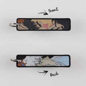 Pode incluir: Um chaveiro preto e branco com uma imagem colorida de duas pessoas na frente e uma única pessoa atrás. O chaveiro é feito de tecido e tem um anel de metal.