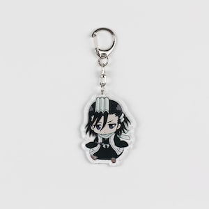 Puede incluir: Un llavero de acr&iacute;lico transparente con un personaje de anime de estilo chibi con cabello negro, una bufanda blanca y un atuendo negro. El personaje tiene una expresi&oacute;n seria y sostiene una espada.