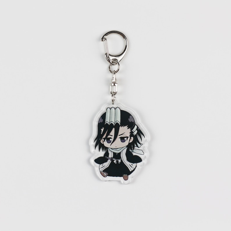 Puede incluir: Un llavero de acr&iacute;lico transparente con un personaje de anime de estilo chibi con cabello negro, una bufanda blanca y un atuendo negro. El personaje tiene una expresi&oacute;n seria y sostiene una espada.