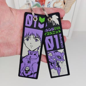 Puede incluir: Dos llaveros rectangulares bordados con ilustraciones de estilo anime. Uno muestra a un personaje de pelo morado con traje morado, el otro un robot mecánico morado. Ambos incluyen el texto 01, caracteres japoneses y logotipos de NERV en negro, blanco y verde.