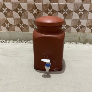 Könnte beinhalten: Ein Terrakotta-Wasserdispenser mit einem weißen Hahn und einem Deckel. Der Dispenser steht auf einer weißen Oberfläche.